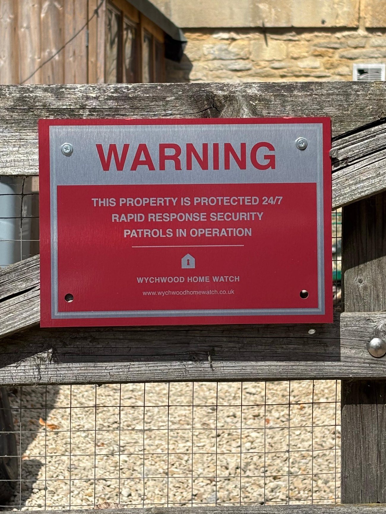 The Wychwood Home Watch warning sign mounted on a gate, with Cotswold stone behind